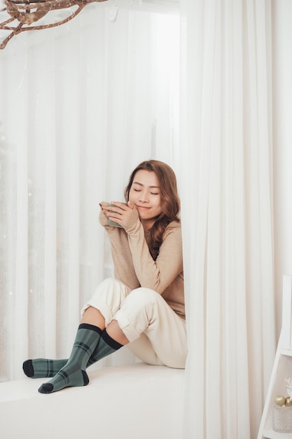 Beautiful woman with curled hair is sitting on the windowsill in a knitted sweater and leg warmers