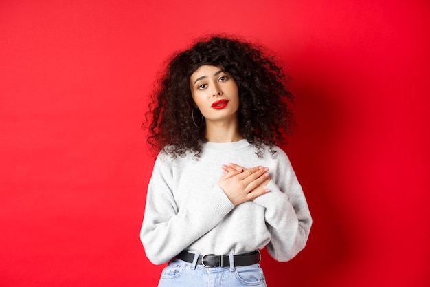 Beautiful woman with curly hair feeling touched and thankful, holding hands on heart and looking with affection at camera, red background