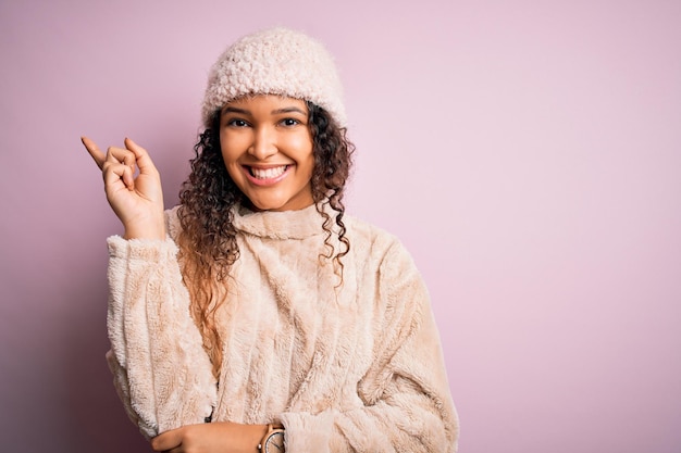 Beautiful woman with curly hair wearing casual sweater and wool cap over pink background with a big smile on face pointing with hand and finger to the side looking at the camera