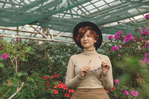 A beautiful woman with eyeglasses and hat smiling among the green plants and flowers.