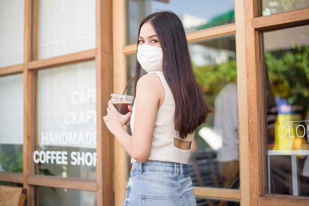 フェイスマスクを持つ美しい女性がコーヒーショップの前でコーヒーを保持しています。