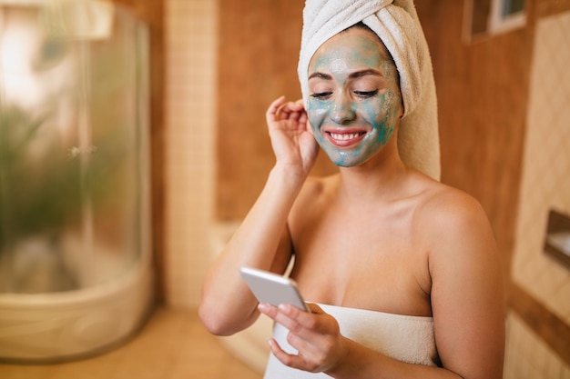 Beautiful woman with facial mask texting on mobile phone in the bathroom