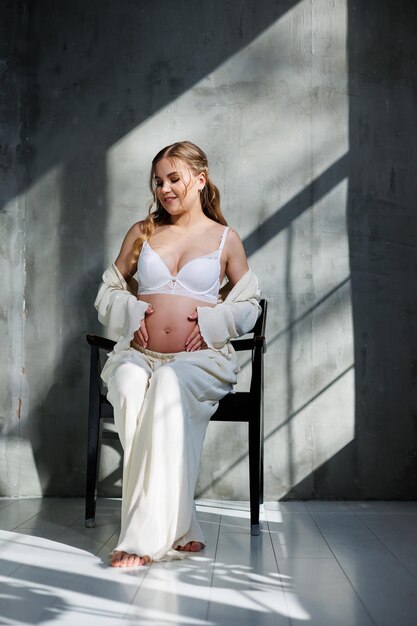 A beautiful woman with fair hair is pregnant in a white elegant suit sitting on a chair on a gray background Fashion for pregnant women A stylish woman is pregnant