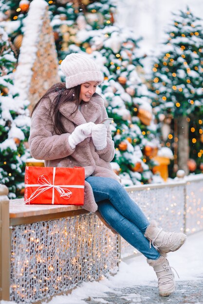 素晴らしい冬の日に屋外の雪の中でクリスマスツリーの近くに贈り物を持つ美しい女性