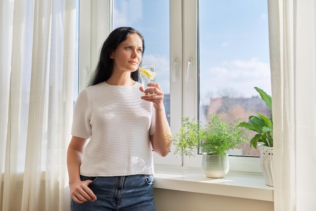 집 창문 근처에 레몬이 든 물 한 잔을 든 아름다운 여성