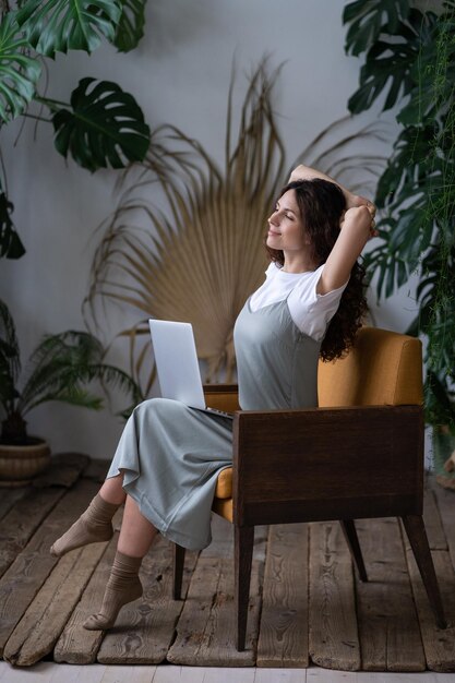 Beautiful woman with hands behind head and computer on knees resting daydreaming in surround plants