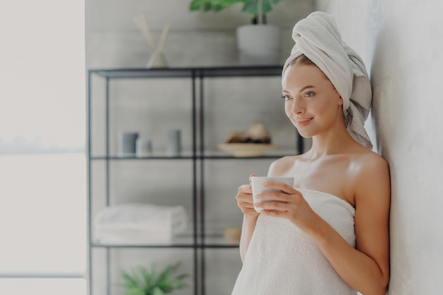 Beautiful woman with healthy skin towel wrapped poses with tea concentrated near wall in cozy home