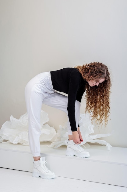 Una bella donna con lunghi capelli ricci in un maglione nero, pantaloni bianchi, scarpe da ginnastica bianche in pelle. nuova collezione di scarpe casual da donna