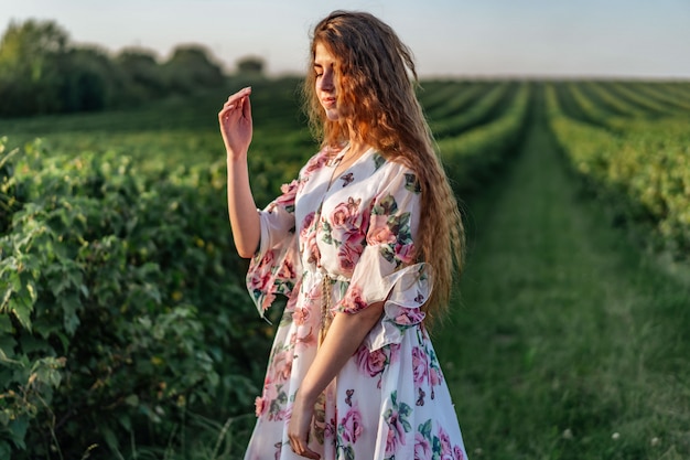 Beautiful woman with long curly hair and freckles face on currant field. woman in a light dress walks in the summer sunny day