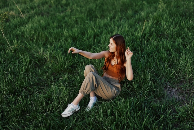 Красивая женщина с длинными вьющимися рыжими волосами сидит летом в парке на зеленой траве в лучах заката