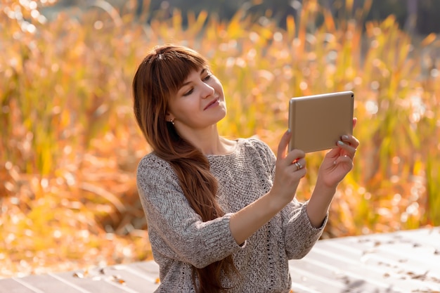 長い髪の美しい女性が屋外の秋の公園に座って、デジタルタブレットを使用してインターネット上で通信します。