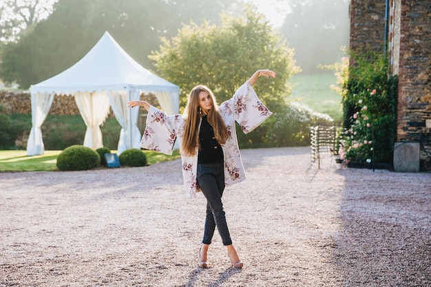 Beautiful woman with long straight hair wears white cape jeans and high heeled shoes stands crossed legs gestures with hands aside poses at camera against ancient citys background charming nature