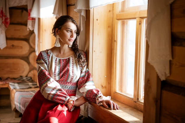 A beautiful woman with a national dress is sitting at the window in the hut