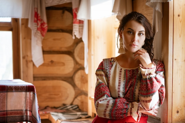 民族衣装を着た美しい女性が小屋の窓に座っています。