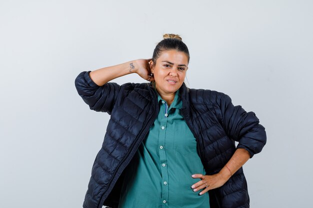 Beautiful woman with one hand behind head, another hand on hip in green shirt, black jacket and looking happy , front view.