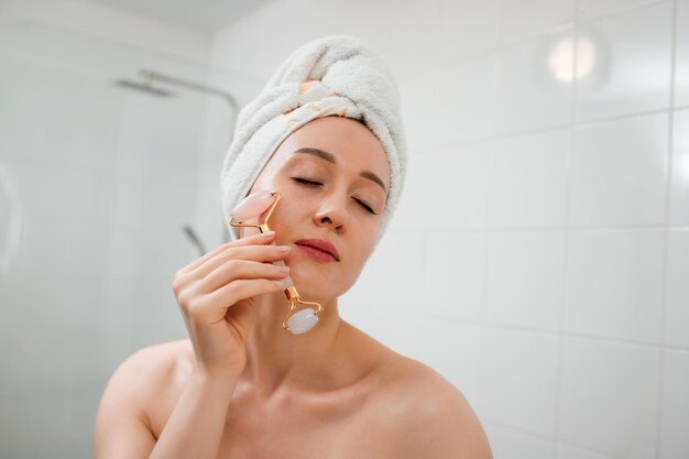 Beautiful woman with perfect skin wearing white towel after
shower making face massage using a jade face roller with natural
quartz stone in bathroom natural cosmetics concept wrinkle
smoothing