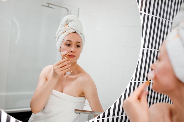 Beautiful woman with perfect skin wearing white towel after\
shower making face massage using a jade face roller with natural\
quartz stone in bathroom natural cosmetics concept wrinkle\
smoothing