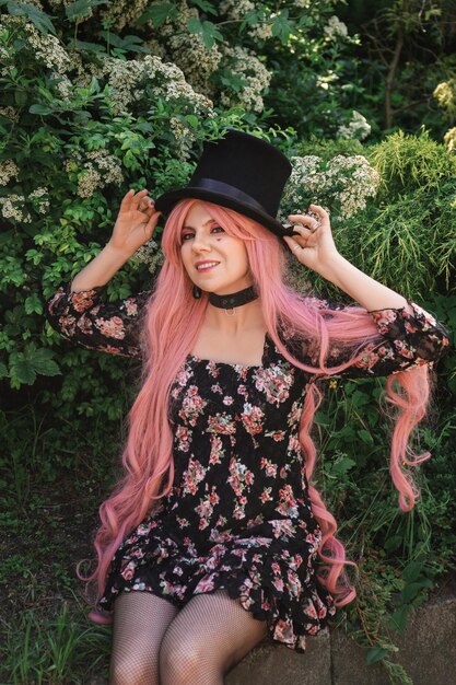 beautiful woman with pink hair in a top hat and in a black dress sitting in the garden in flowers