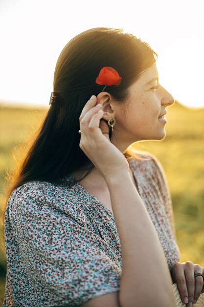 Beautiful woman with poppy flower behind ear relaxing in barley field in sunset light Stylish female standing in evening summer countryside Atmospheric moment rustic slow life