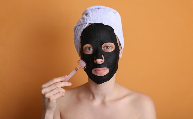 Beautiful woman with purifying black mask on her face and towel on head Girl massages her face with massage roller Orange background Facial skin care