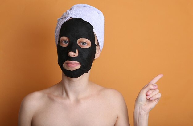 Beautiful woman with purifying black mask on her face and towel on head Girl shows polish on copy space Orange background