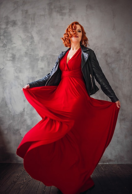 Photo beautiful woman with red hair and in red dress with leather jacket poses before a grey wall