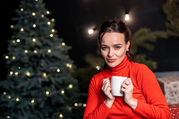 Beautiful woman with red lipstick sitting and holding a white cup in her hands Girl with a white mug near the Christmas tree decorated with lights