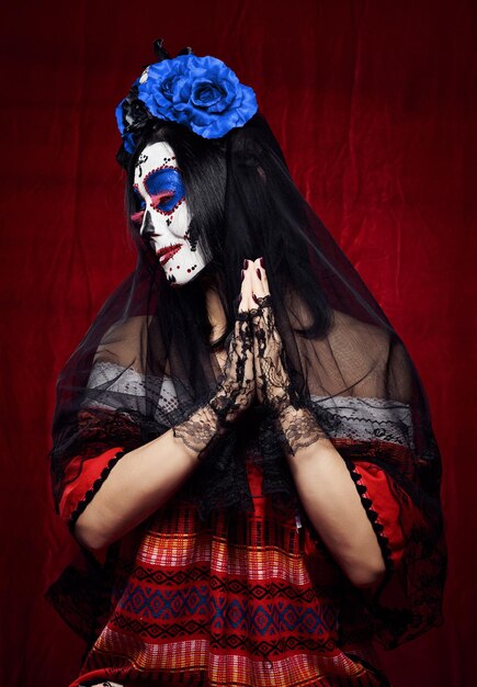 Beautiful woman with a sugar skull makeup with a wreath of flowers on her head and a skull hands raised up in prayer pose in black gloves, red background