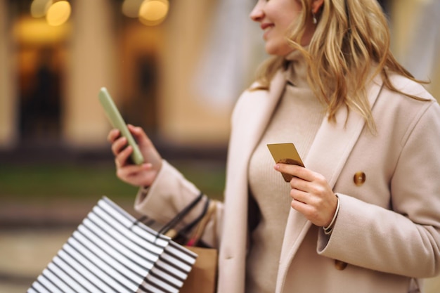 Foto bella donna con carta di credito telefonica, ragazza bionda in cappotto che tiene i sacchetti della spesa sulla strada della città