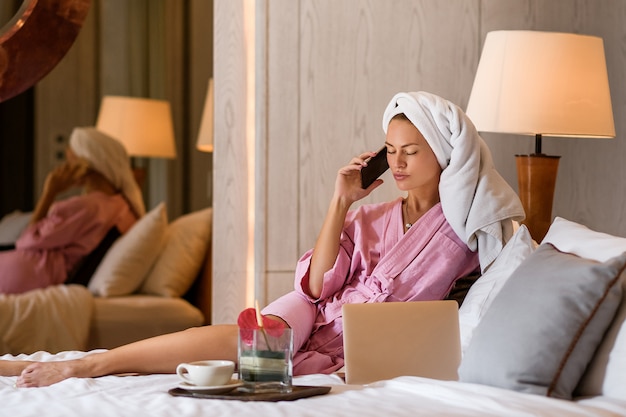 Bella donna con l'asciugamano sulla testa che parla sul telefono in camera da letto a casa. la routine mattutina.