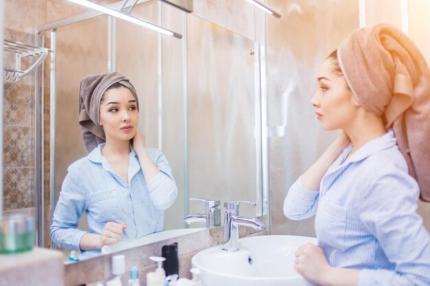 Beautiful woman with a towel on her head, looking in the mirror in the bathroom