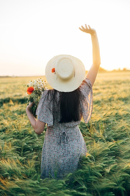 Beautiful woman with wildflowers enjoying sunset in barley field Atmospheric tranquil moment rustic slow life Stylish female gathering flowers and relaxing in evening summer countryside