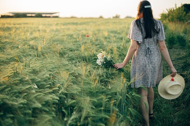야생 꽃과  모자를 입은 아름다운 여인이 해가 지는 빛에 보리에서 걸어 가고 있으며, 멋진 여인이 저녁에 여름의 시골에서 휴식을 취하고 꽃을 모으고, 분위기가 조용한 순간입니다.