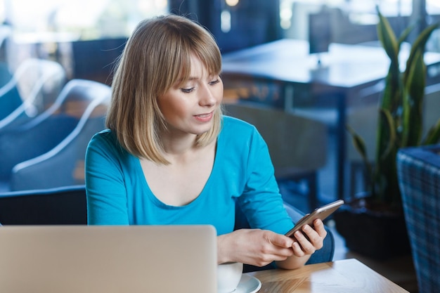 Beautiful woman working on laptop having break using cell phone checking social networks