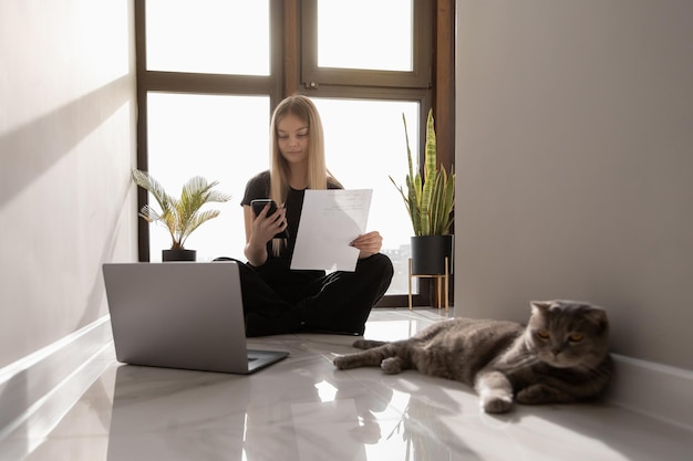 Красивая женщина работает дома за компьютером, сидя на полу у окна и проверяет документы и уведомления по телефону