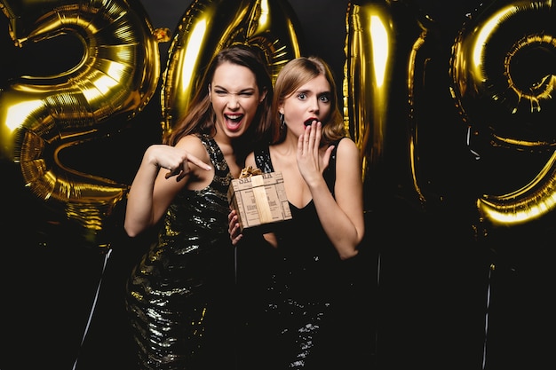 Beautiful Women Celebrating New Year. Happy Gorgeous Girls In Stylish Sexy Party Dresses Holding Gold 2019 Balloons, Having Fun At New Year's Eve Party. Holiday Celebration. High Quality Image