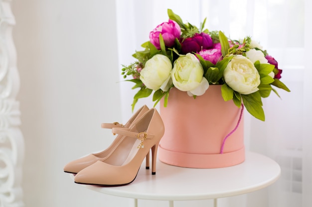 Beautiful women's beige high-heeled shoes stand on a white table, next to a round box with colorful peonies