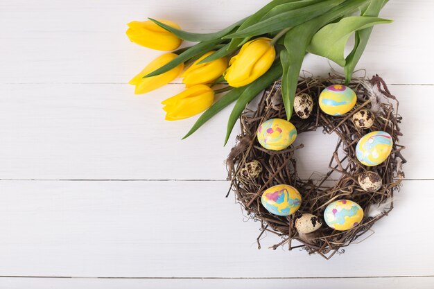 Beautiful yellow tulips with colorful quail and chicken eggs in wreath on white wooden table. Spring and Easter holiday concept.