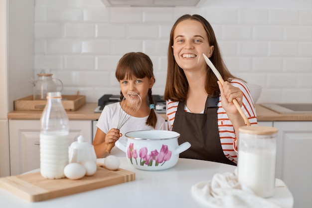 Beautiful young adult mom and her cute little daughter playing and smiling while baking in kitchen at home looking at camera with happy smiling