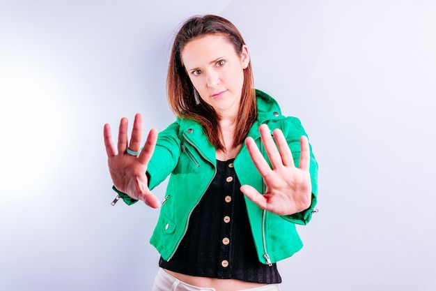 Beautiful young adult woman in green jacket showing palms in\
front with serious gesture with white background with copyspace.\
concept send away distance