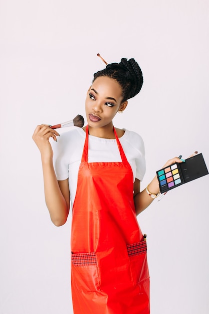 Beautiful young African American beautician woman holding set of make up brushes and eye-shadows, isolated on white