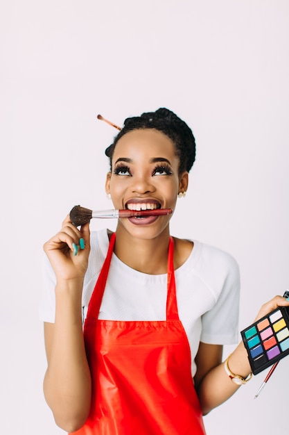 Beautiful young African American beautician woman holding set of make up brushes and eye-shadows, isolated on white