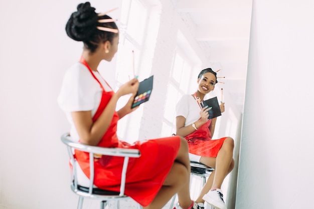メイクアップブラシのセットを保持していると鏡で見ている美しい若いアフリカ系アメリカ人美容師女性