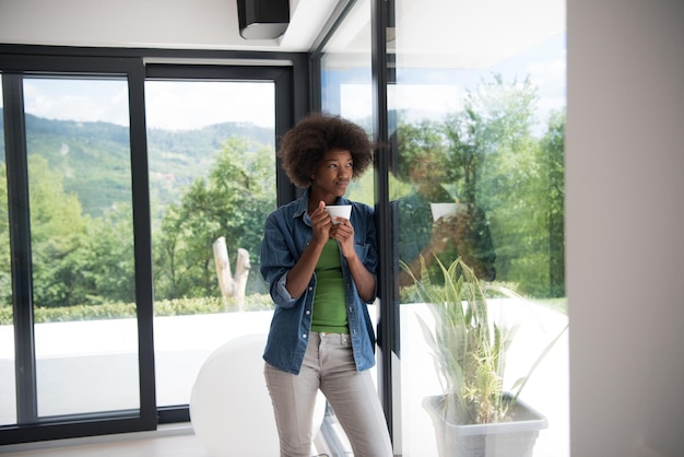 Bella giovane donna afroamericana che beve caffè e guarda attraverso una finestra nella sua casa di lusso