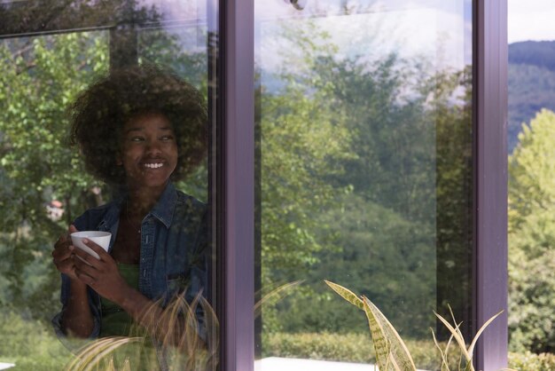 Beautiful young african american woman drinking coffee and looking through a window in her luxury home