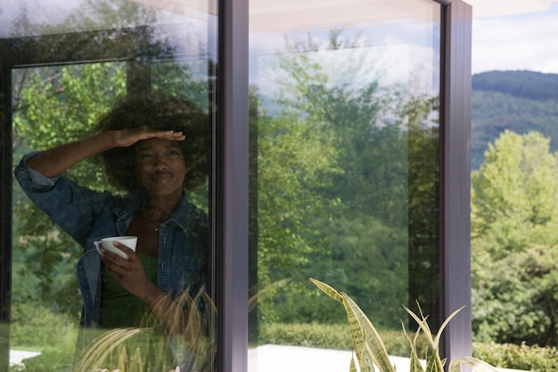 Beautiful young african american woman drinking coffee and looking through a window in her luxury home