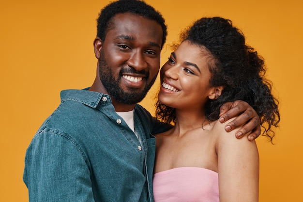 黄色の背景に立っている間抱き締めて笑っている美しい若いアフリカのカップル