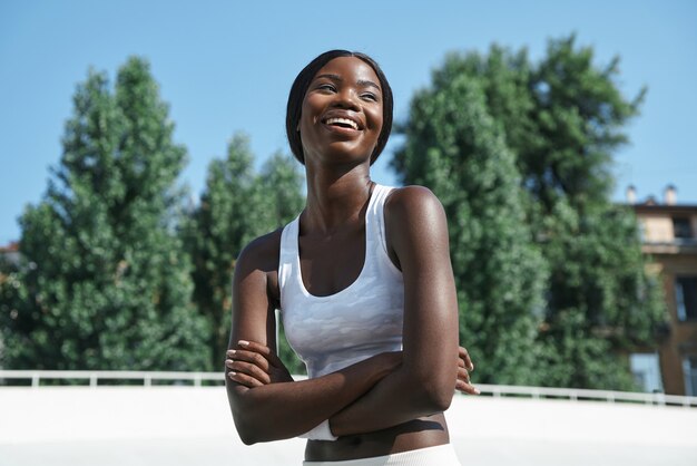 Beautiful young african woman in sports clothing keeping arms crossed and smiling while standing out...