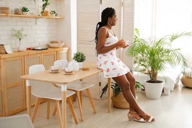 beautiful young african woman in a summer dress in the kitchen drinking tea