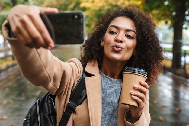 Beautiful young african woman wearing coat walking outdoors at the park, carrying backpack, taking a selfie